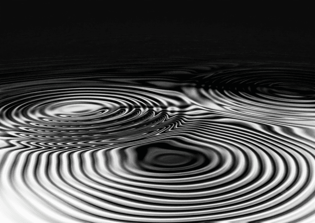Black and white photograph of two circular ripple patterns colliding on a water surface, creating an interference pattern where the waves cancel and distort each other.